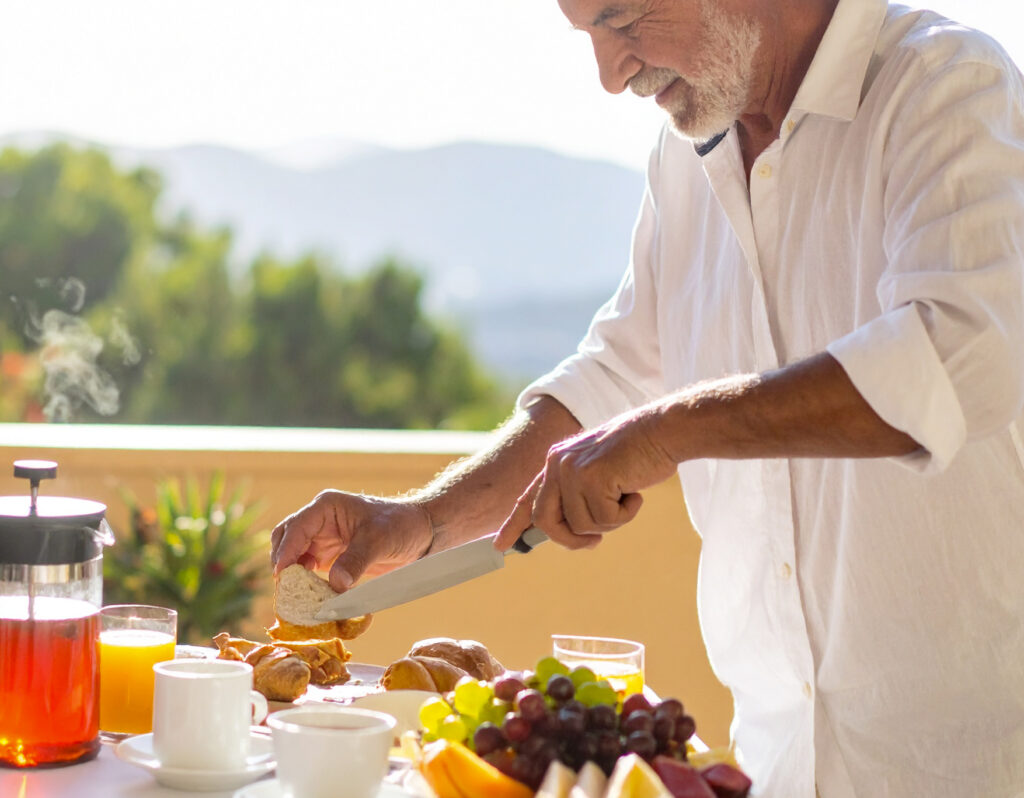 Un-homme-de-55-ans,-en-terrasse,-coupe-une-tartine-à-l’aide-d’un-petit-outil-ergonomi-23278 Homme préparant un petit déjeuner ensoleillé.